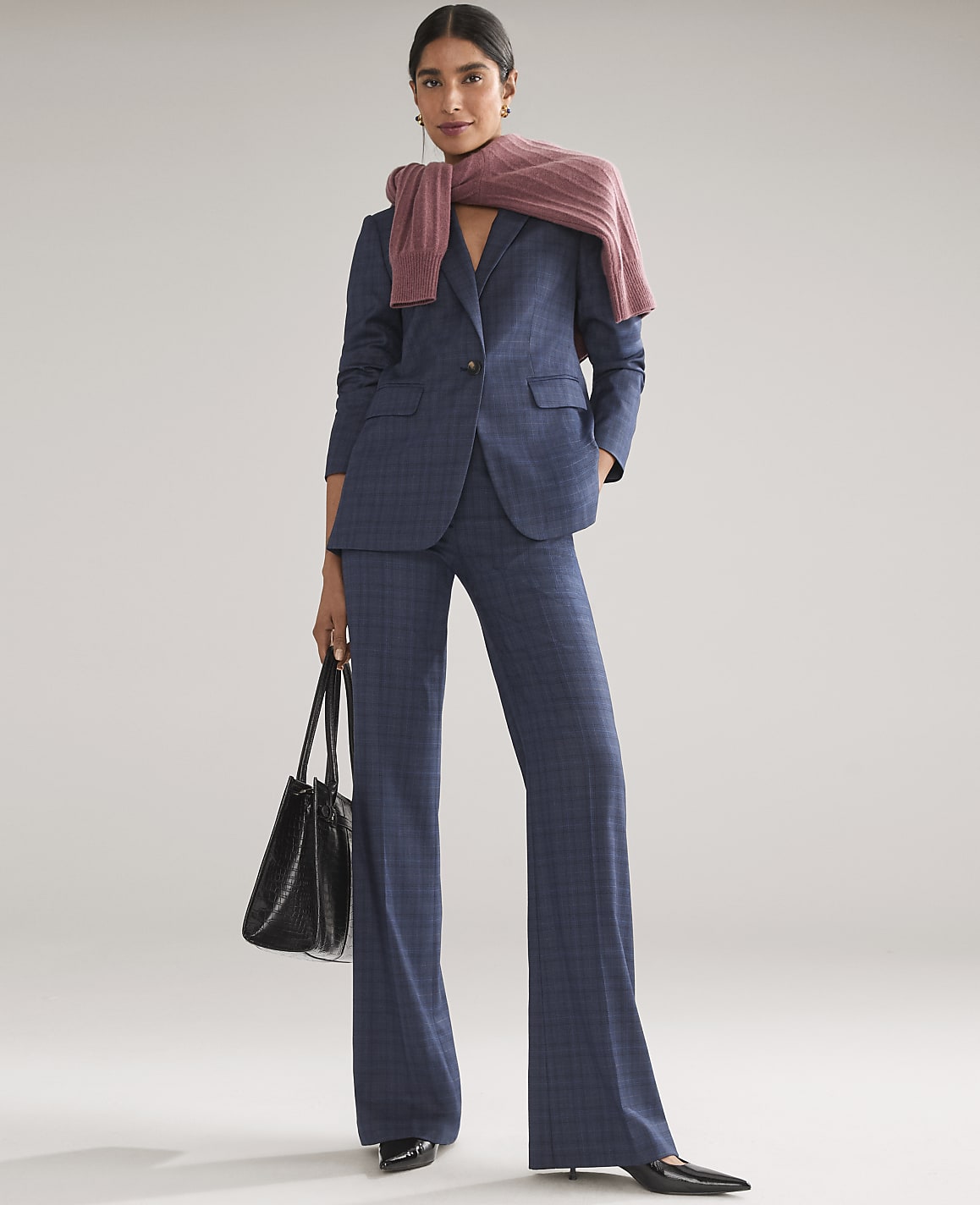 A woman wearing a blue plaid suit with a pink sweater draped over her shoulders and holding a black handbag stands against a plain light gray background.