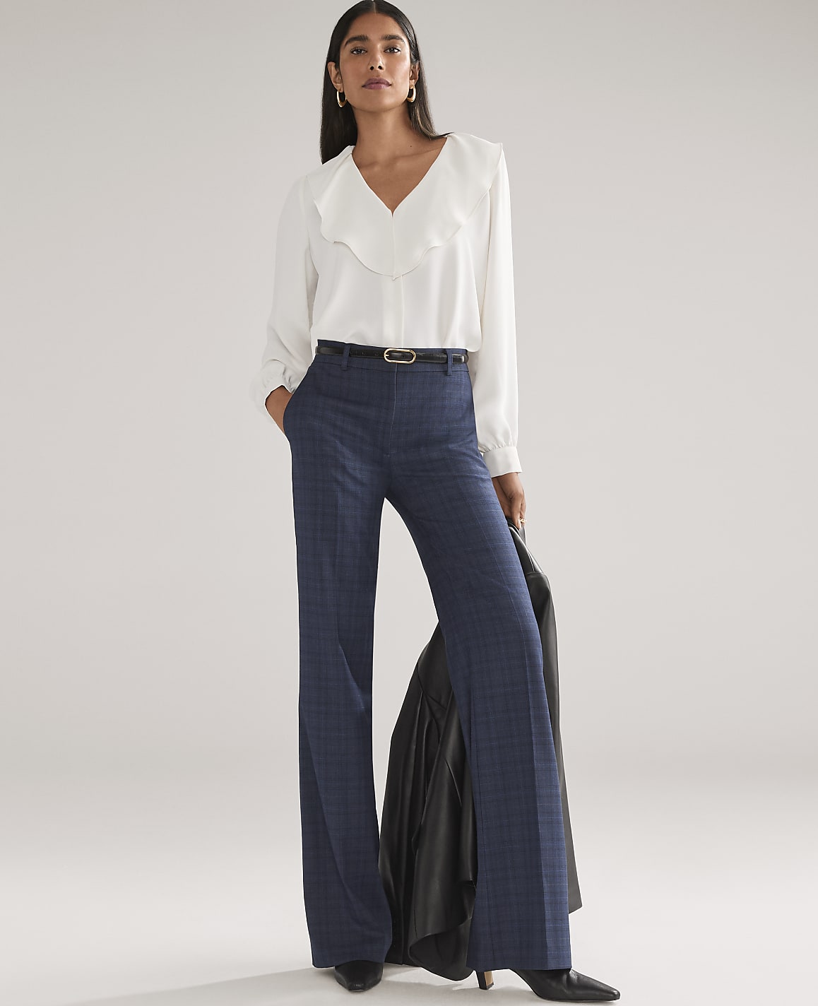 A woman wearing a white ruffled blouse and navy blue wide-leg trousers stands against a plain, light grey background.