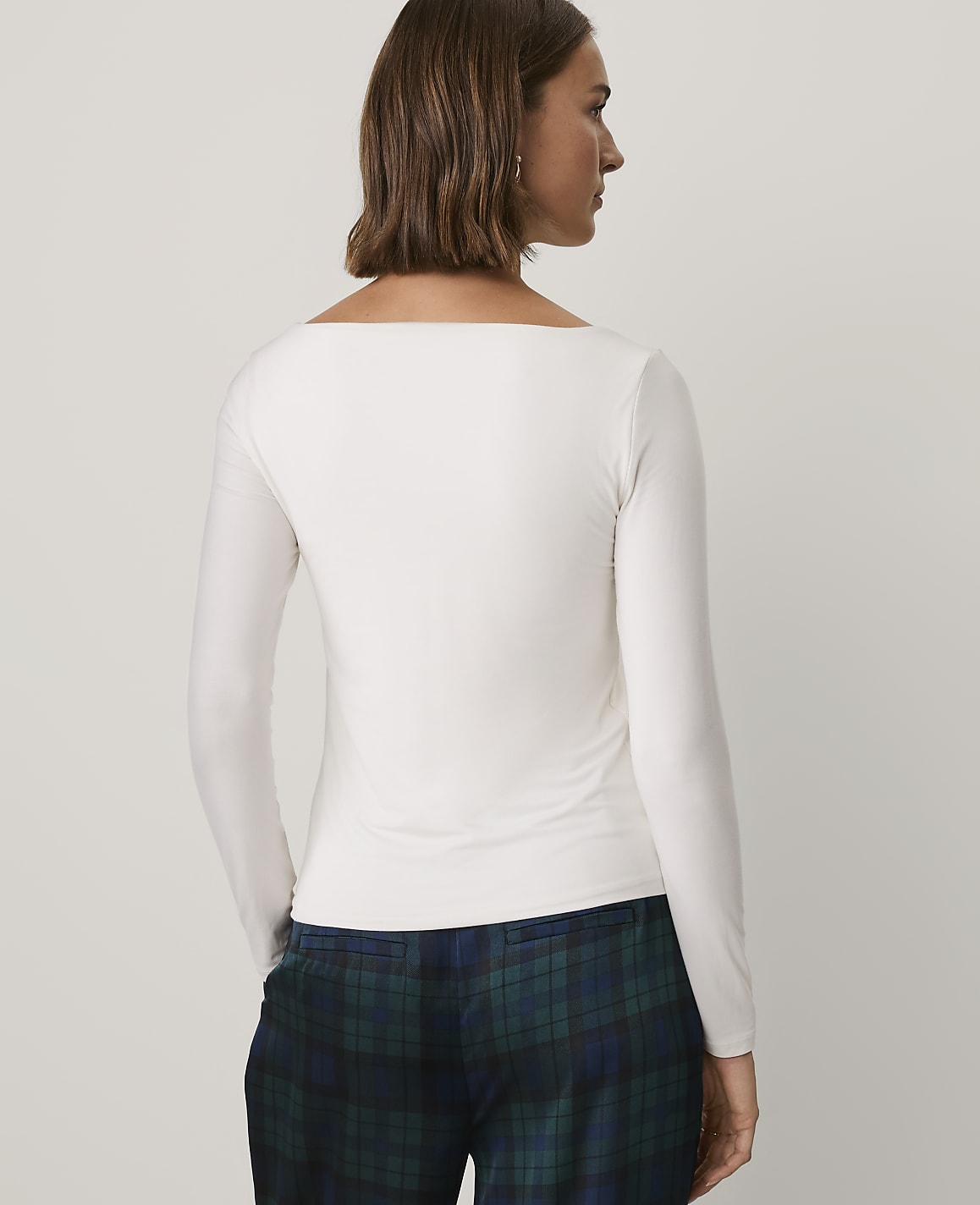 A woman wearing a white long-sleeved top and blue plaid trousers stands against a plain light gray background.