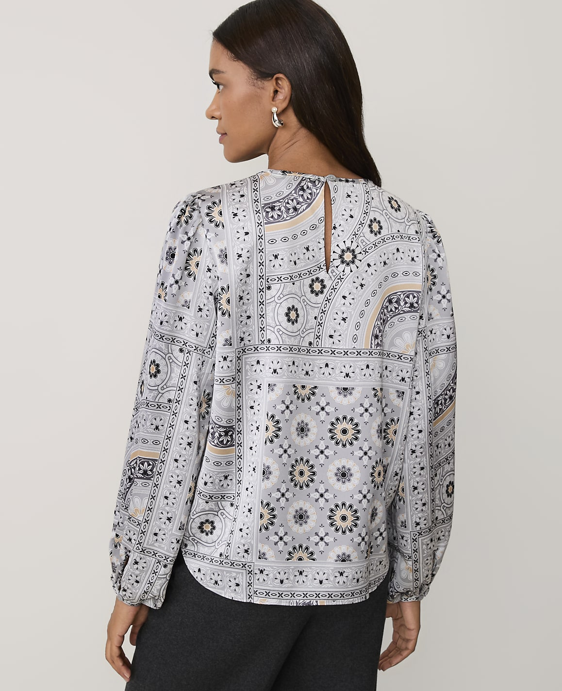 A woman wearing a patterned grey and white blouse and dark trousers stands against a plain light-colored background.