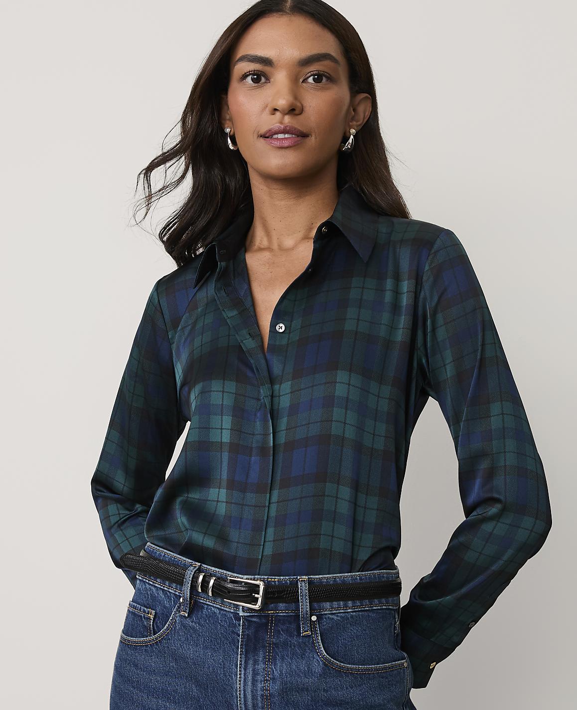 A woman wearing a dark green plaid button-down shirt and blue jeans stands against a plain, light-colored background.