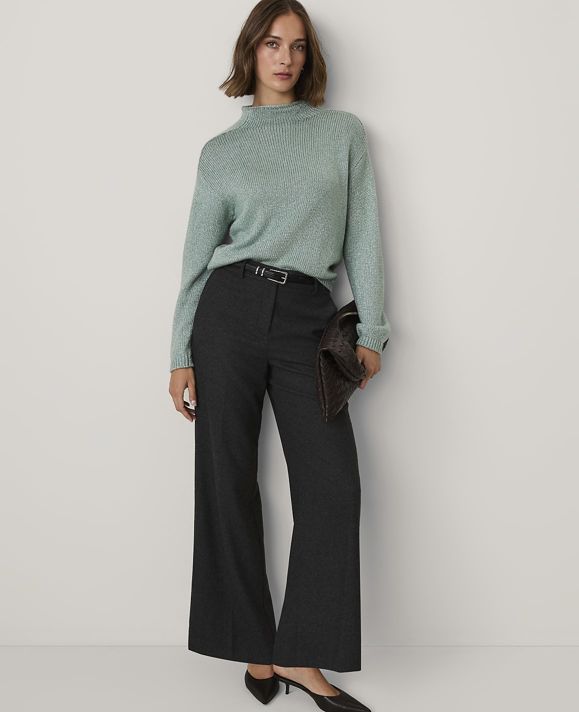 A woman wearing a light green sweater and dark wide-leg trousers stands against a plain white background.