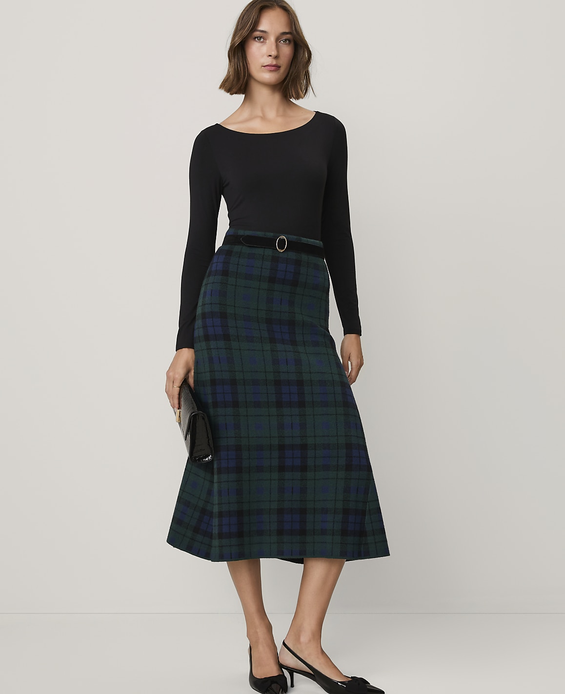A woman wearing a black long-sleeved top and a green plaid midi skirt stands against a plain, light-colored background.