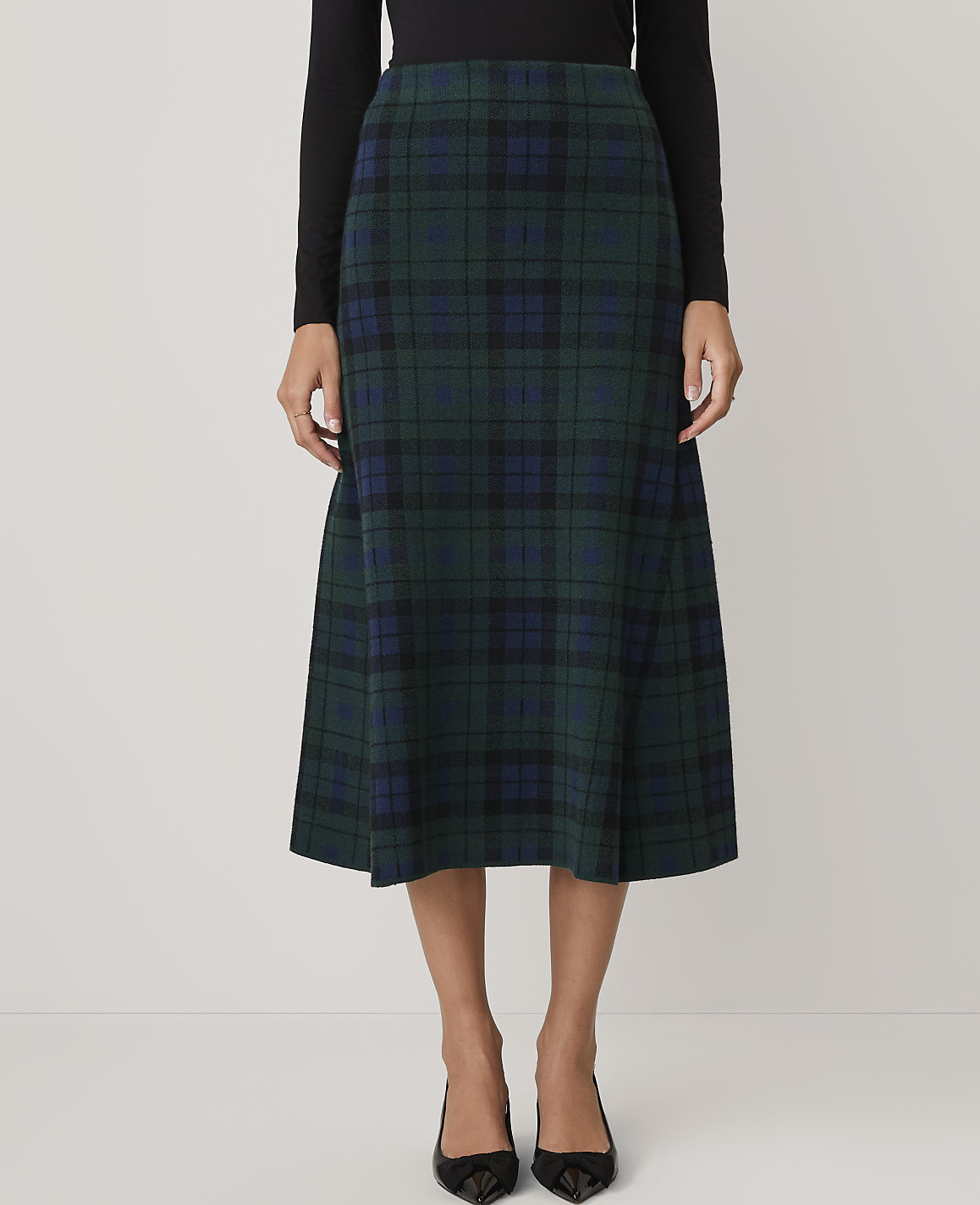 A person wears a black top and a green and blue plaid midi skirt against a plain white background.