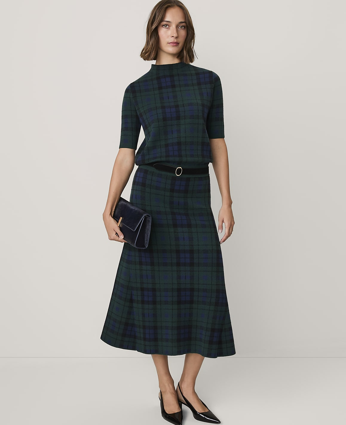 A woman wearing a green plaid two-piece set and black heels stands against a plain white background.