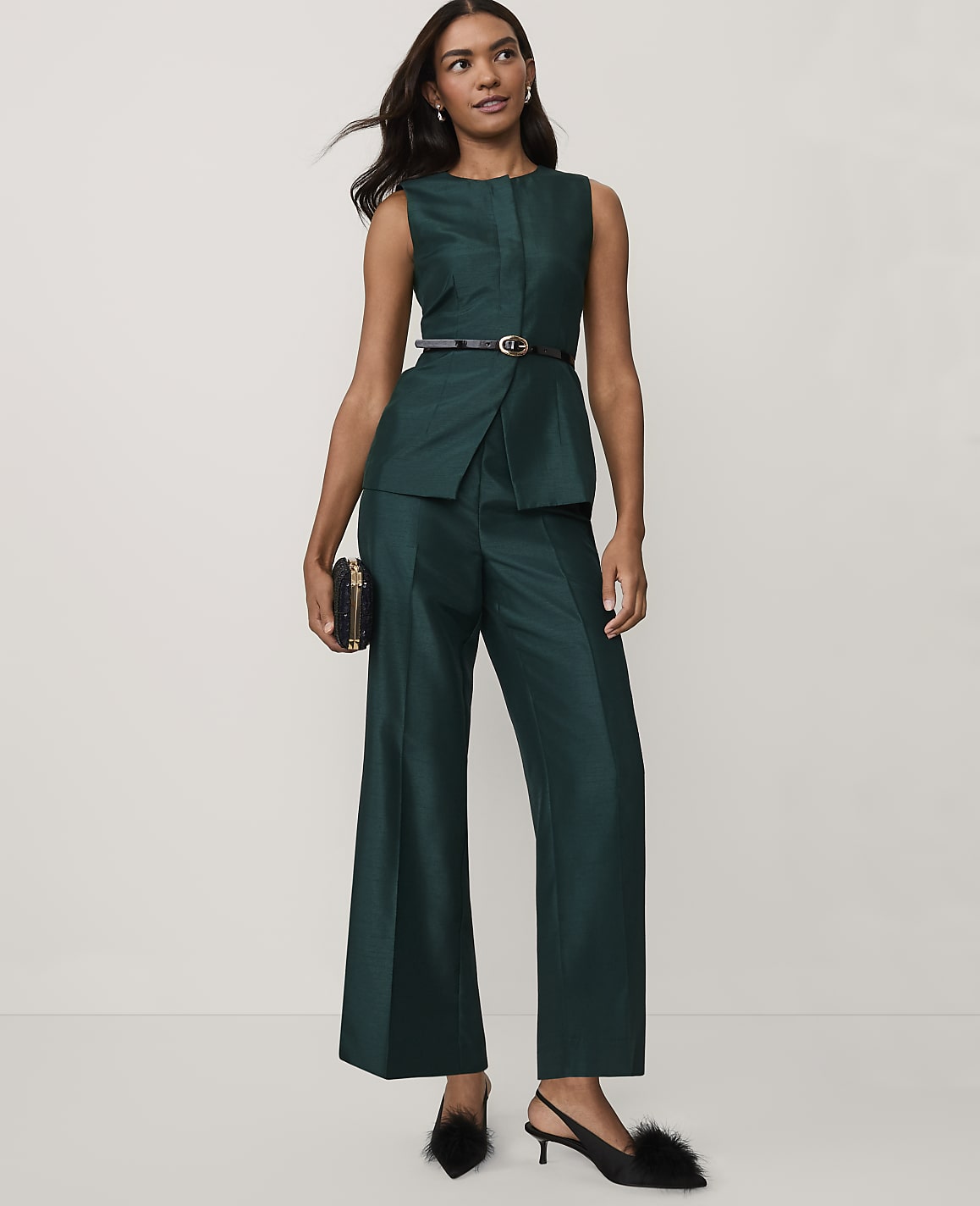 A woman wearing a dark green sleeveless suit and black heels stands against a plain white background.