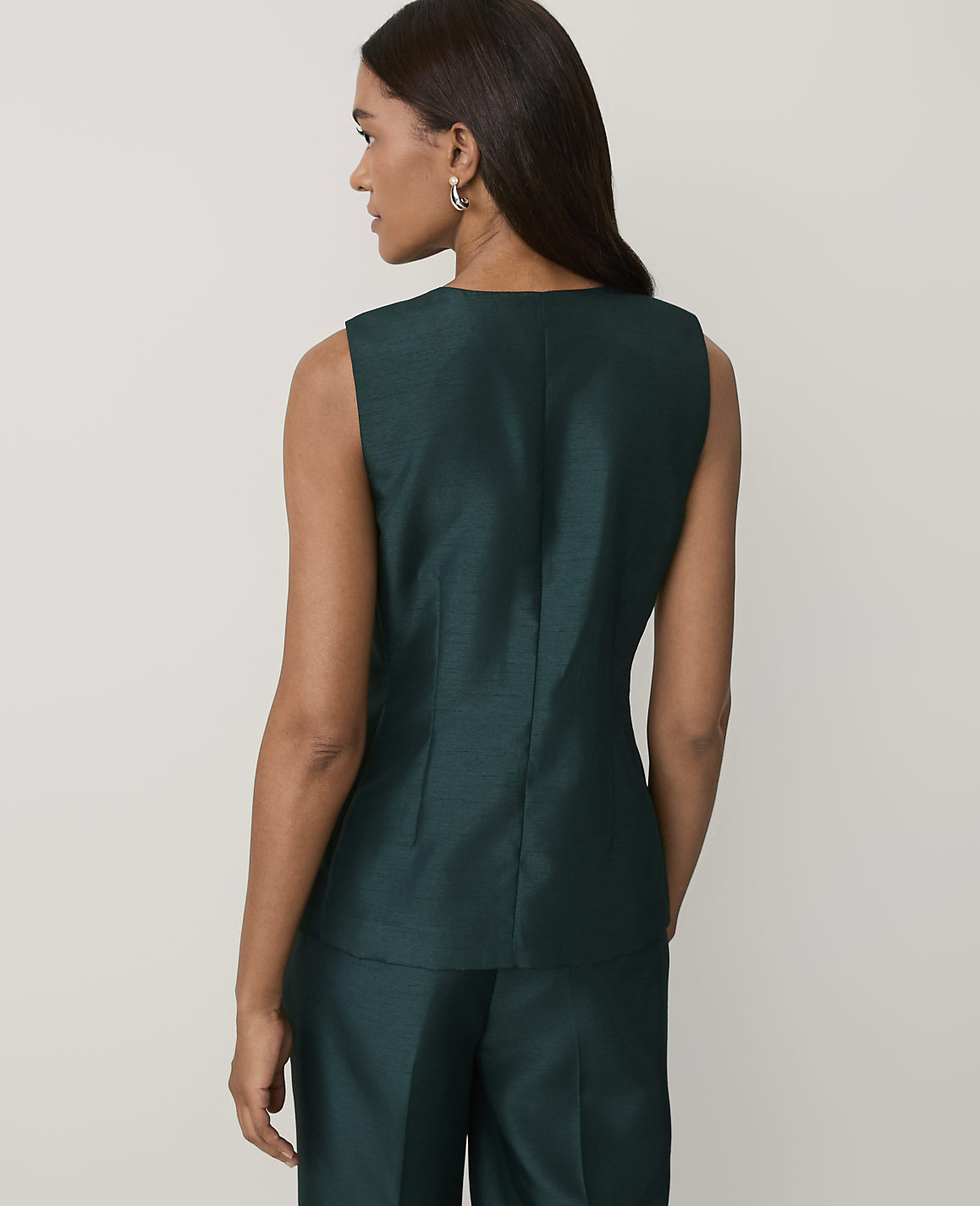 A woman wearing a dark green sleeveless top and matching trousers stands with her back to the camera against a plain light gray background.