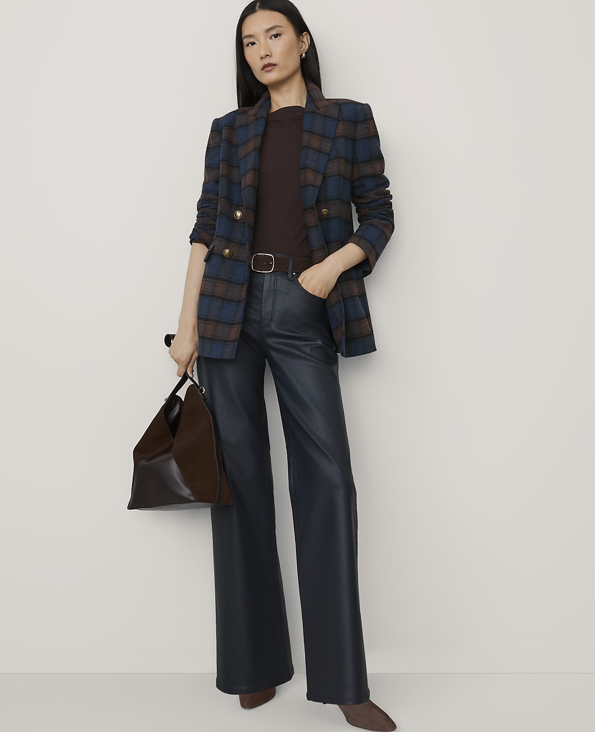 A woman wearing a plaid blazer, dark top, and wide-leg faux leather trousers stands against a plain, light-colored background.