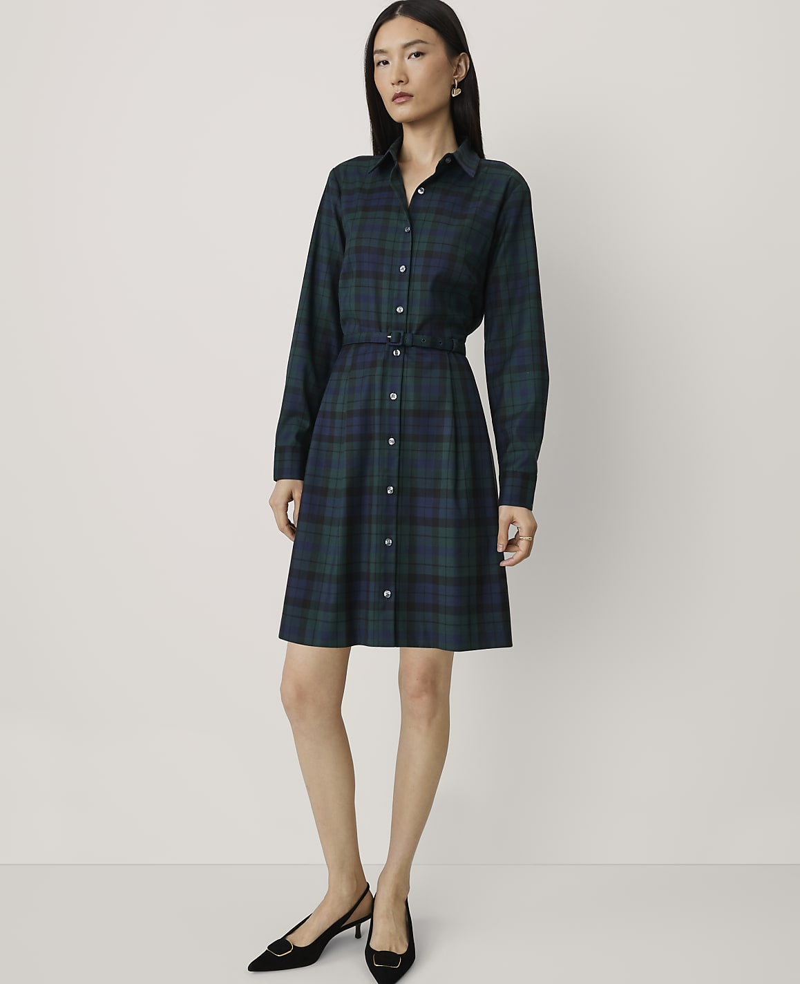 A woman wearing a dark green plaid shirt dress and black heels stands against a plain, light-colored background.