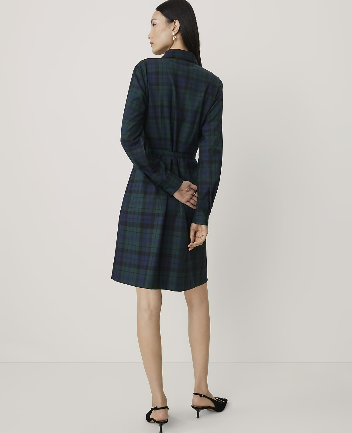 A woman wearing a green plaid shirt dress and black heels stands with her back to the camera against a plain white background.