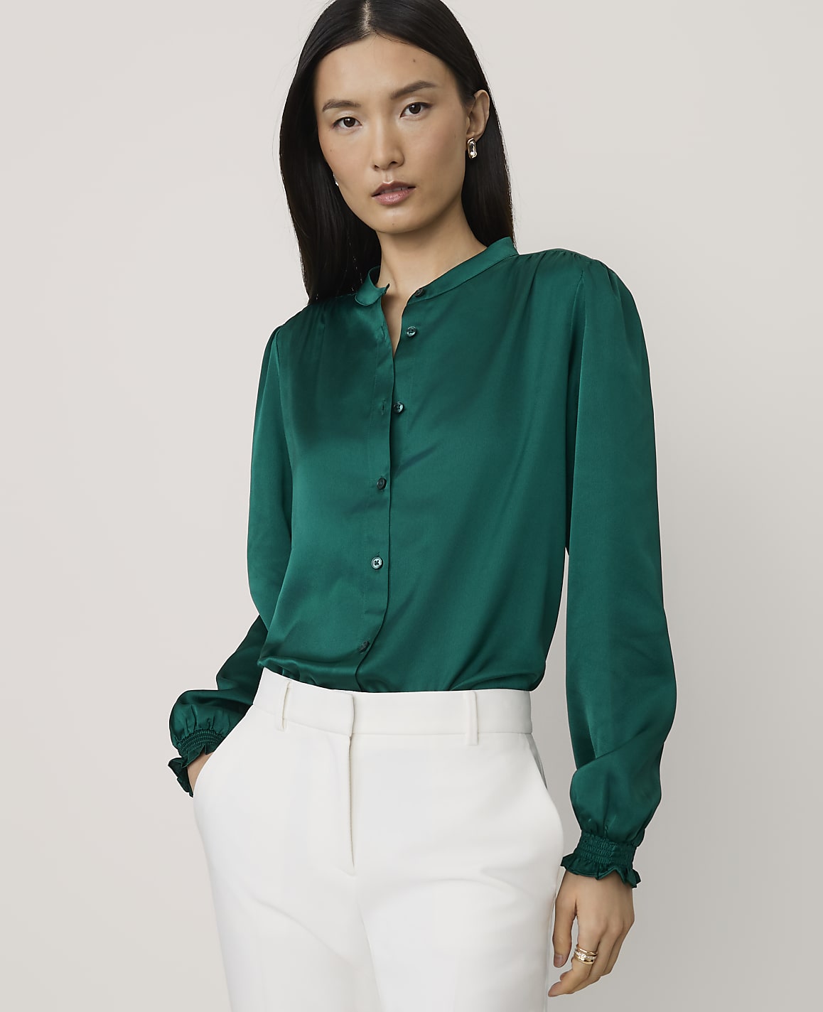 A woman wearing a green satin blouse and white trousers stands against a plain, light-colored background.