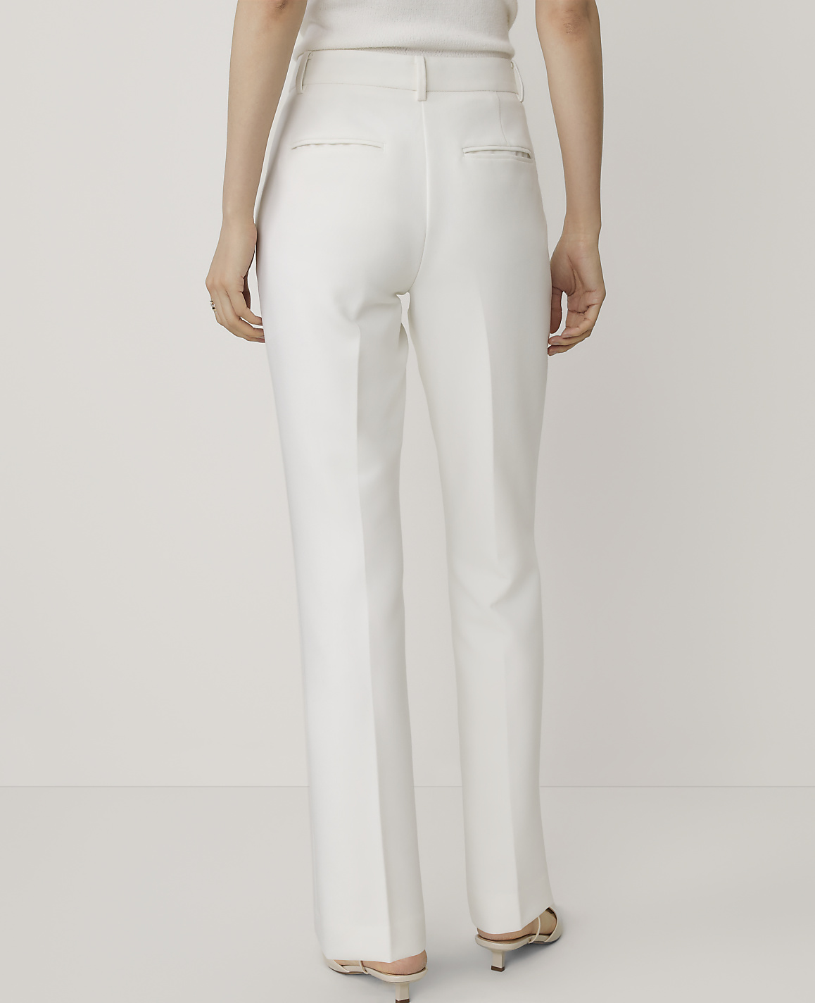 A person stands facing away from the camera wearing white trousers and heels against a plain white background.