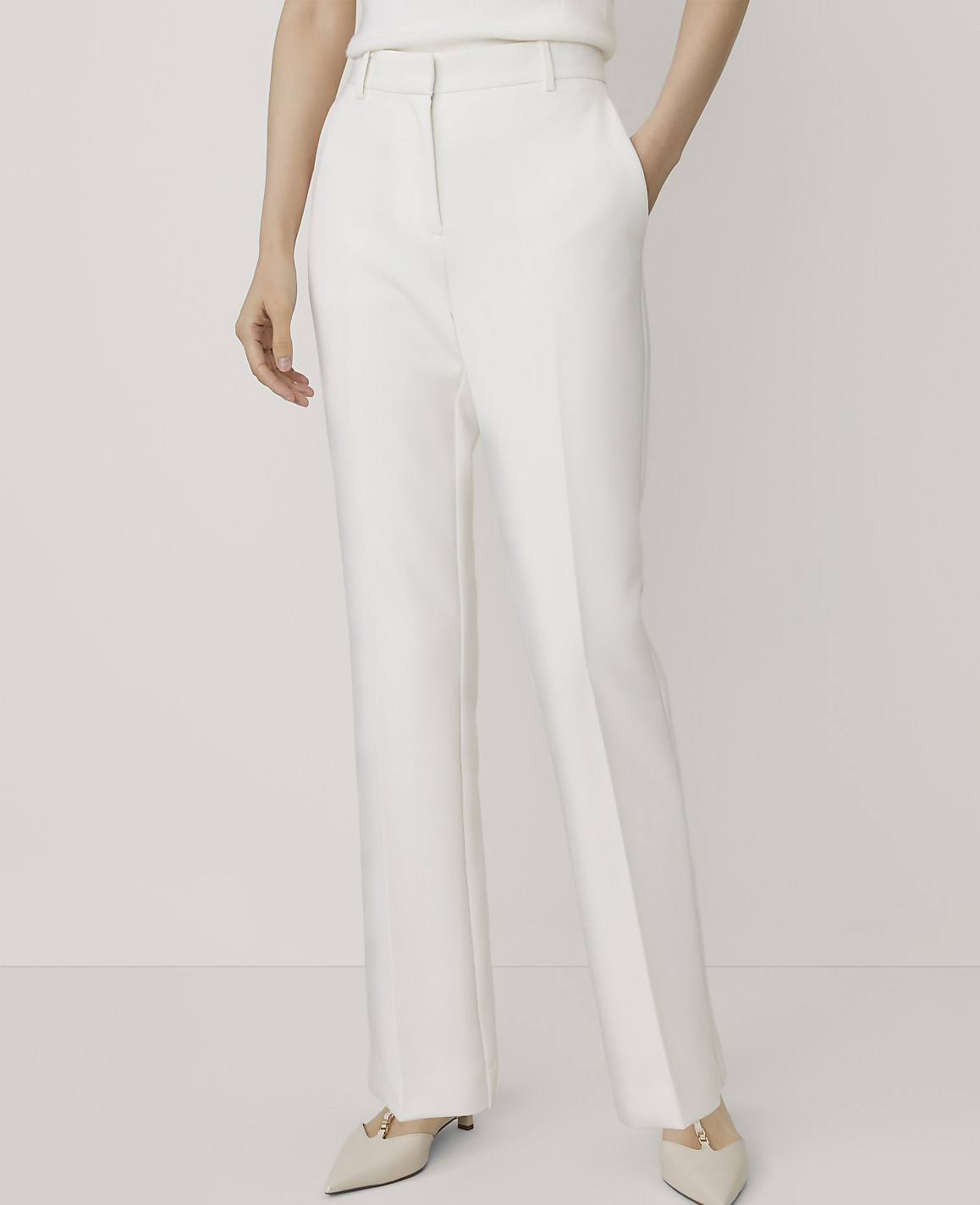 A person wearing white wide-leg trousers and beige pointed-toe heels stands against a plain, light-colored background.