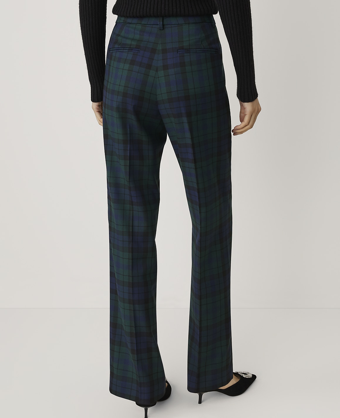 A person wearing a black sweater and green plaid trousers stands against a plain white background.