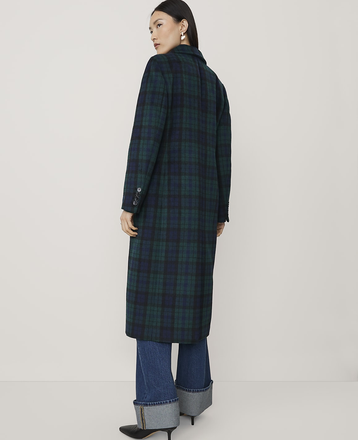 A woman wearing a long green and blue plaid coat and cuffed blue jeans stands against a plain, light-colored background.