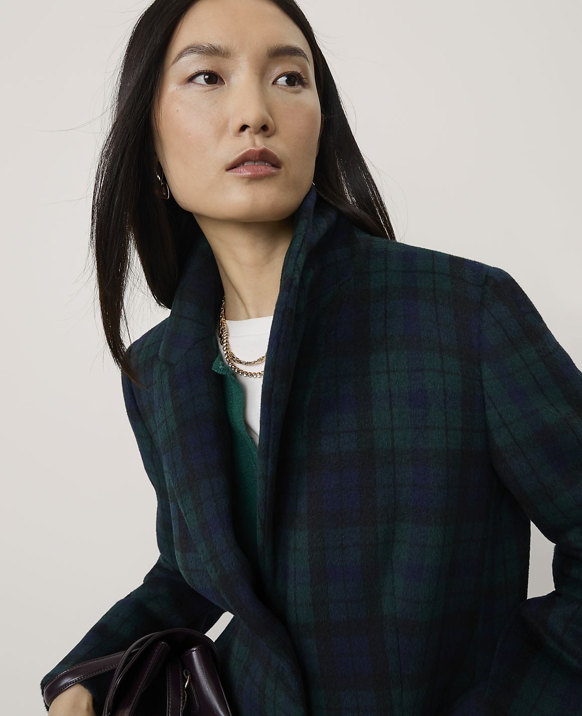 A woman wearing a dark green plaid blazer and a gold chain necklace poses against a plain, light-colored background.
