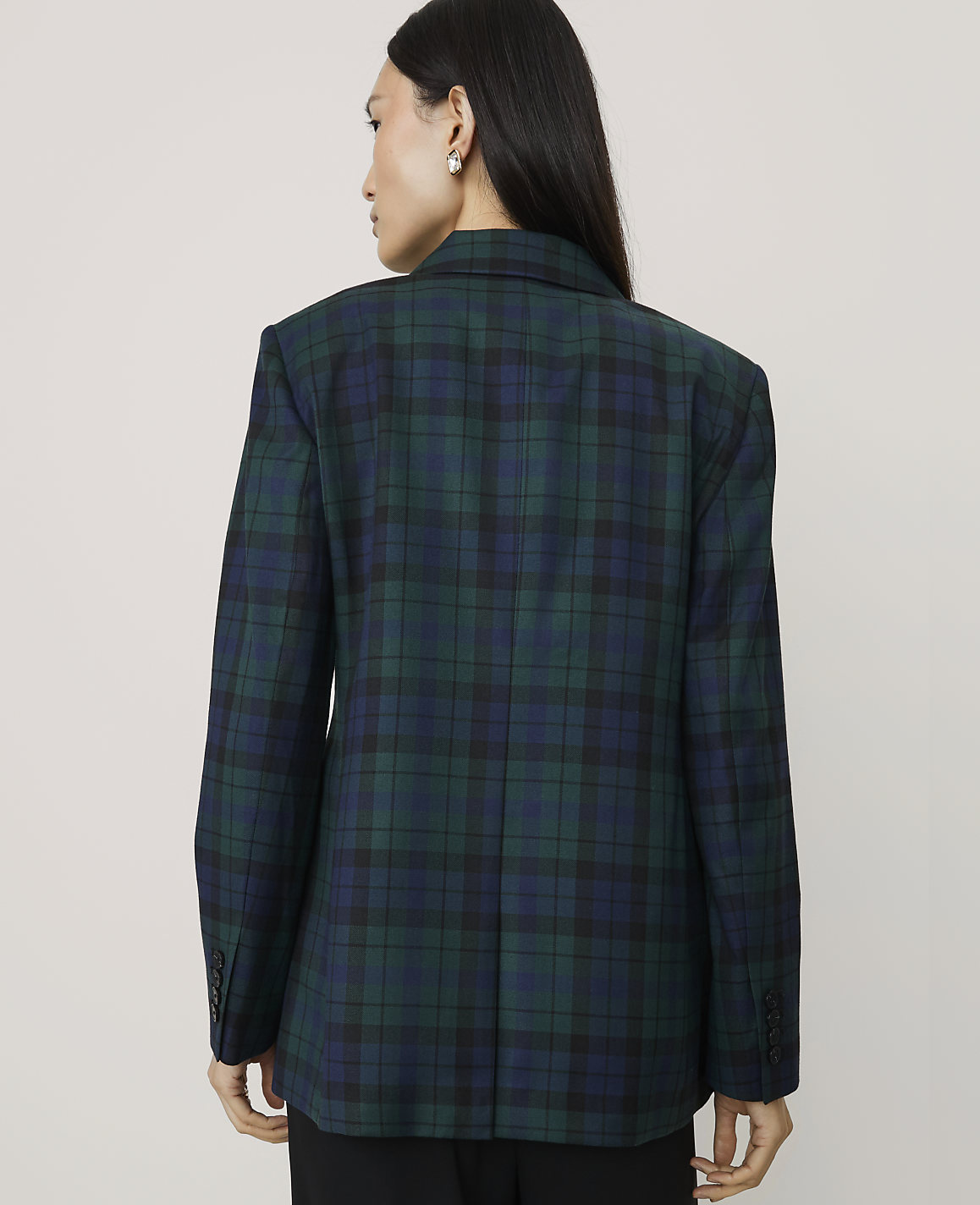 A woman wearing a green and blue plaid blazer stands with her back to the camera against a plain, light-colored background.