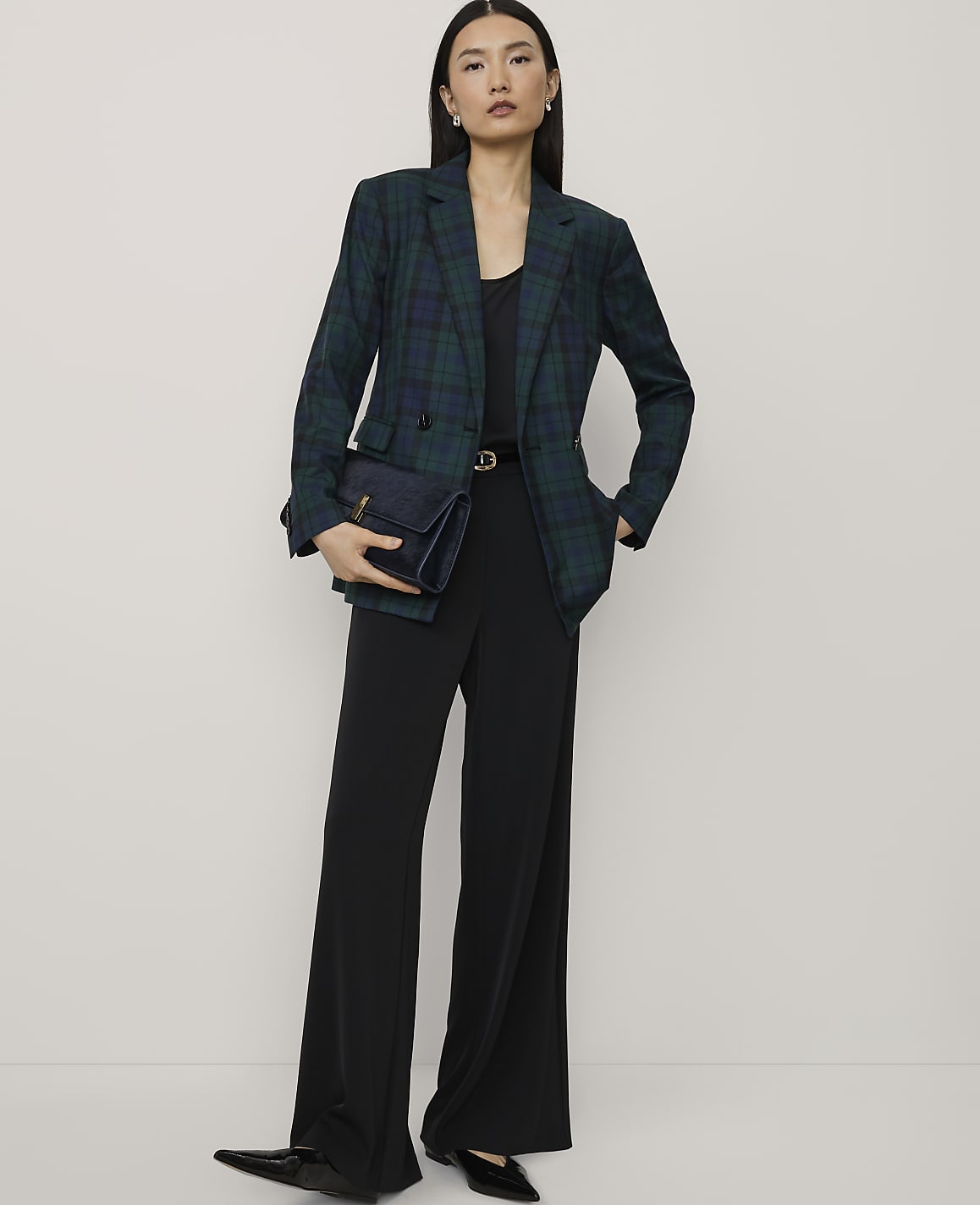 A woman wearing a green plaid blazer and black wide-leg trousers stands against a plain, light-colored background.