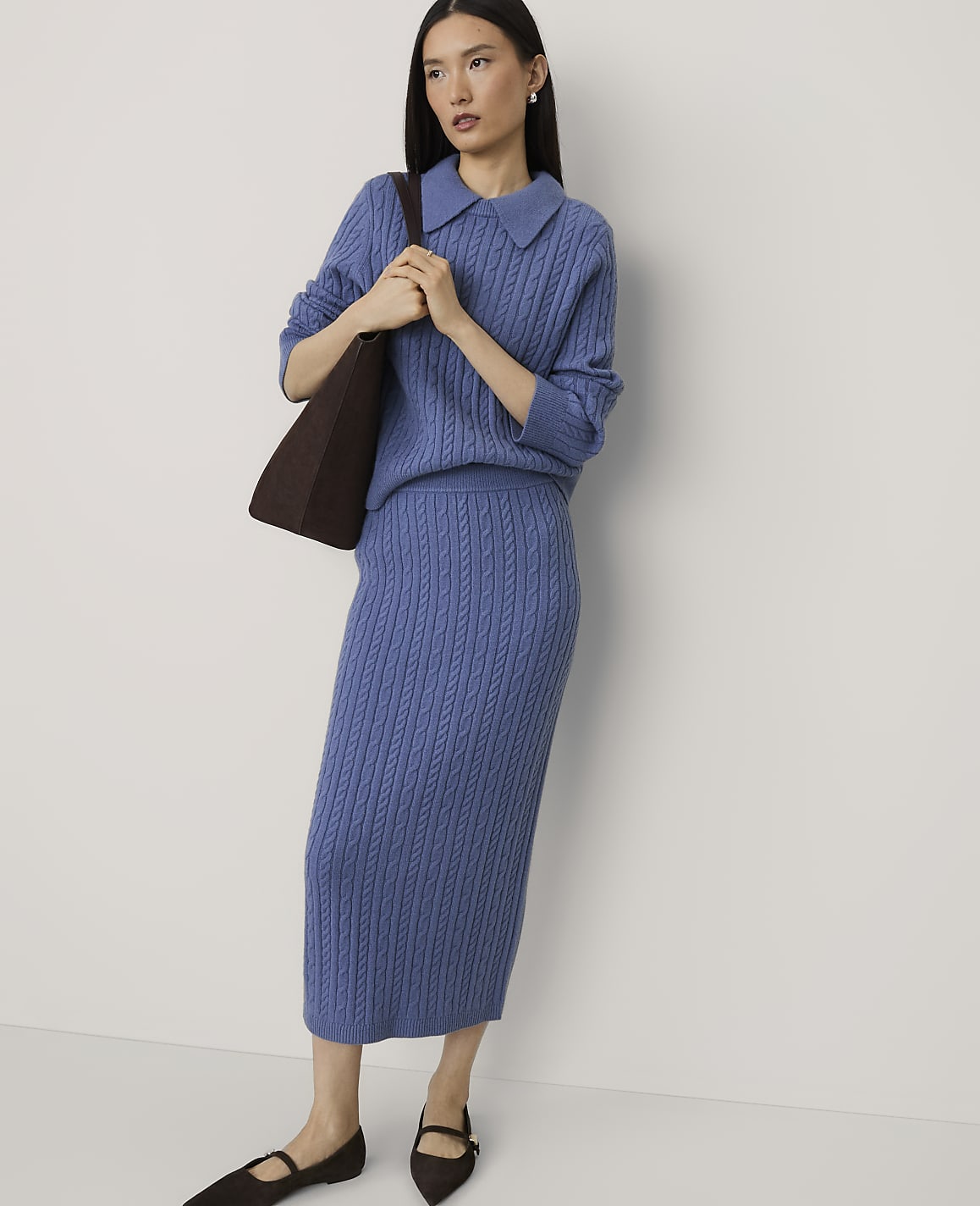 A woman wearing a blue cable-knit sweater and matching skirt stands against a plain white background.