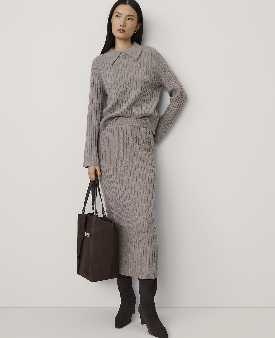 A woman wearing a grey cable-knit sweater and skirt set with dark brown boots and a handbag stands against a plain white wall.