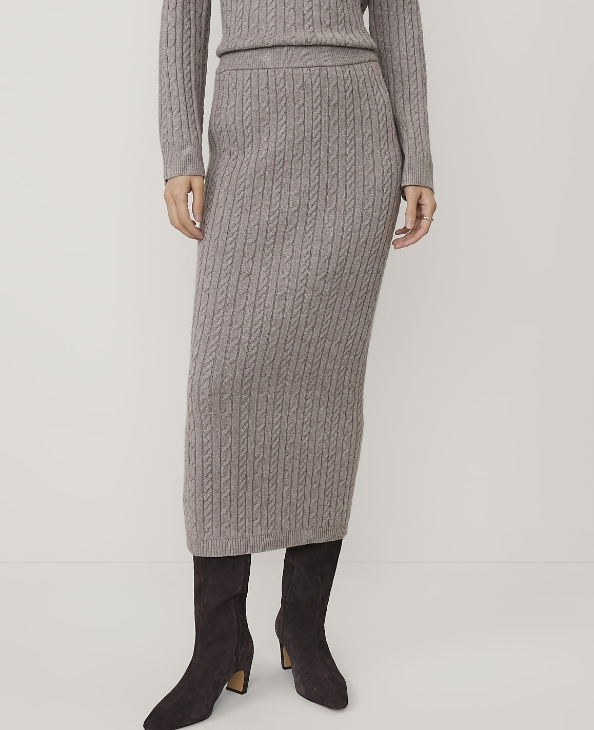 A person wearing a grey cable-knit sweater and matching midi skirt with dark brown ankle boots stands against a plain white background.