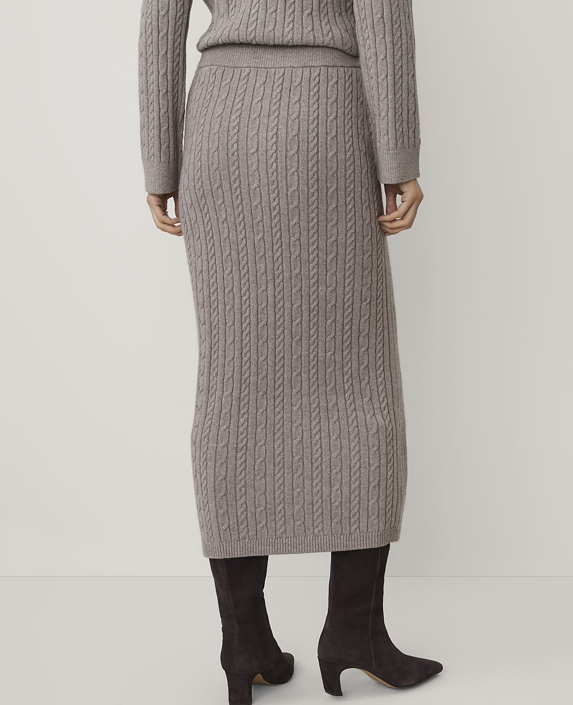 A person stands with their back to the camera wearing a taupe cable-knit sweater and matching skirt with dark brown boots against a plain white background.