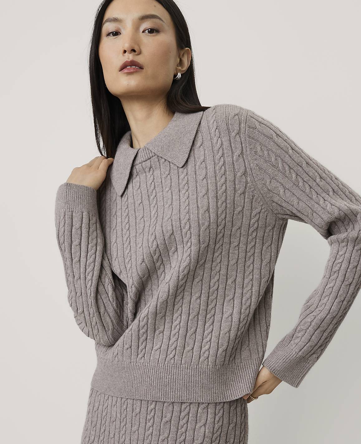 A woman wearing a mauve cable-knit sweater and matching skirt poses against a plain, light-colored background.
