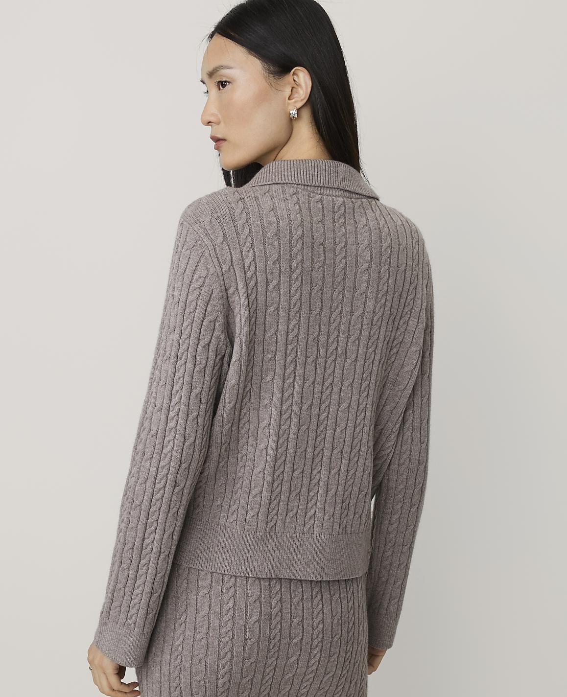 A woman wearing a taupe cable-knit sweater and matching pants stands against a plain, light-colored background.