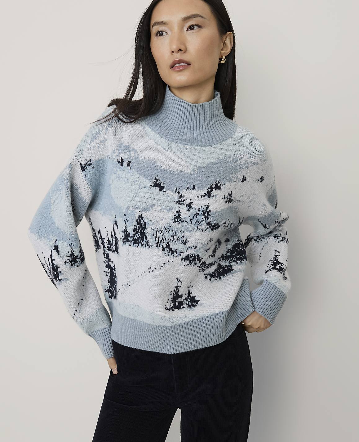 A woman wearing a light blue winter landscape patterned sweater and black pants stands against a plain white background.