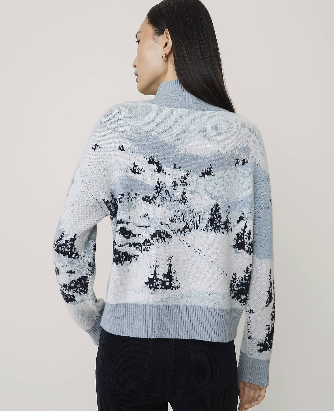 A woman wearing a light blue landscape-patterned sweater and black pants stands against a plain, light-colored background.