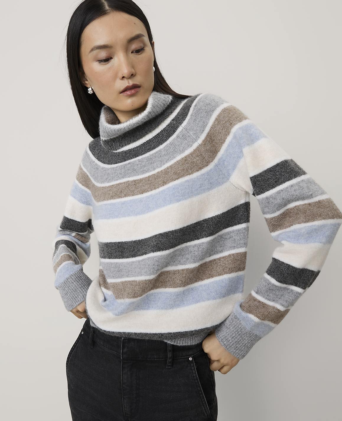A woman wearing a multi-colored striped turtleneck sweater and black pants stands against a plain, light-colored background.