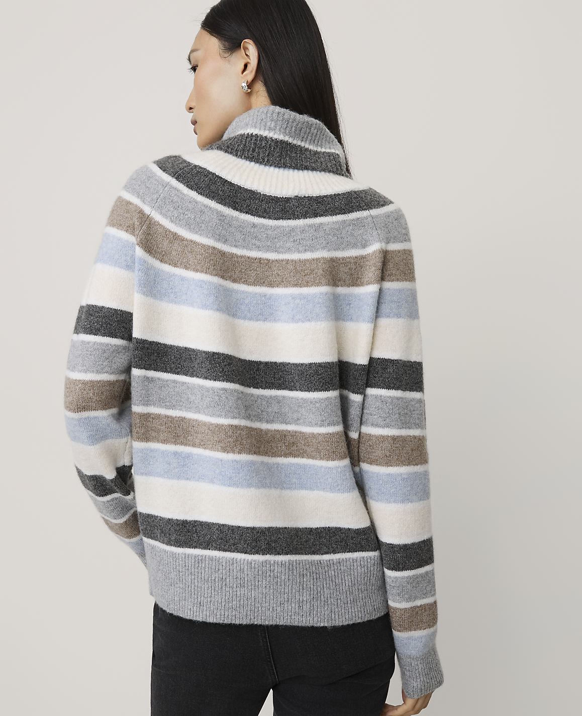 A woman wearing a multi-colored striped sweater and black pants stands against a plain, light-colored background.