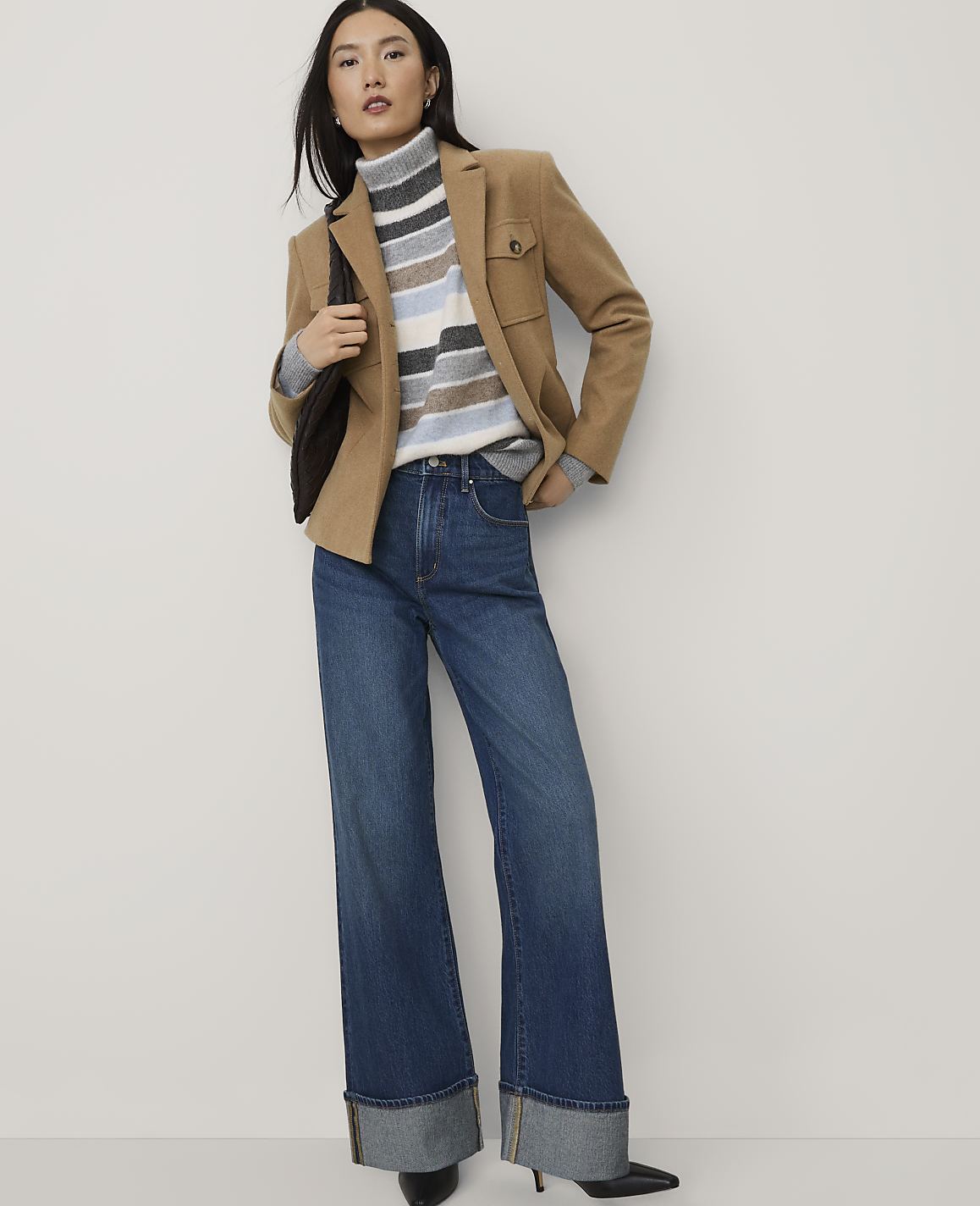 A woman wearing a tan blazer, a striped turtleneck, and wide-leg jeans stands against a plain white background.