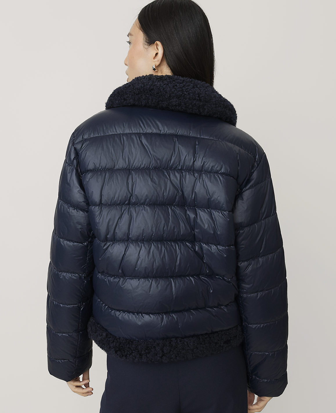 A woman wearing a navy blue puffer jacket with a faux-fur collar stands against a plain, light-colored background.
