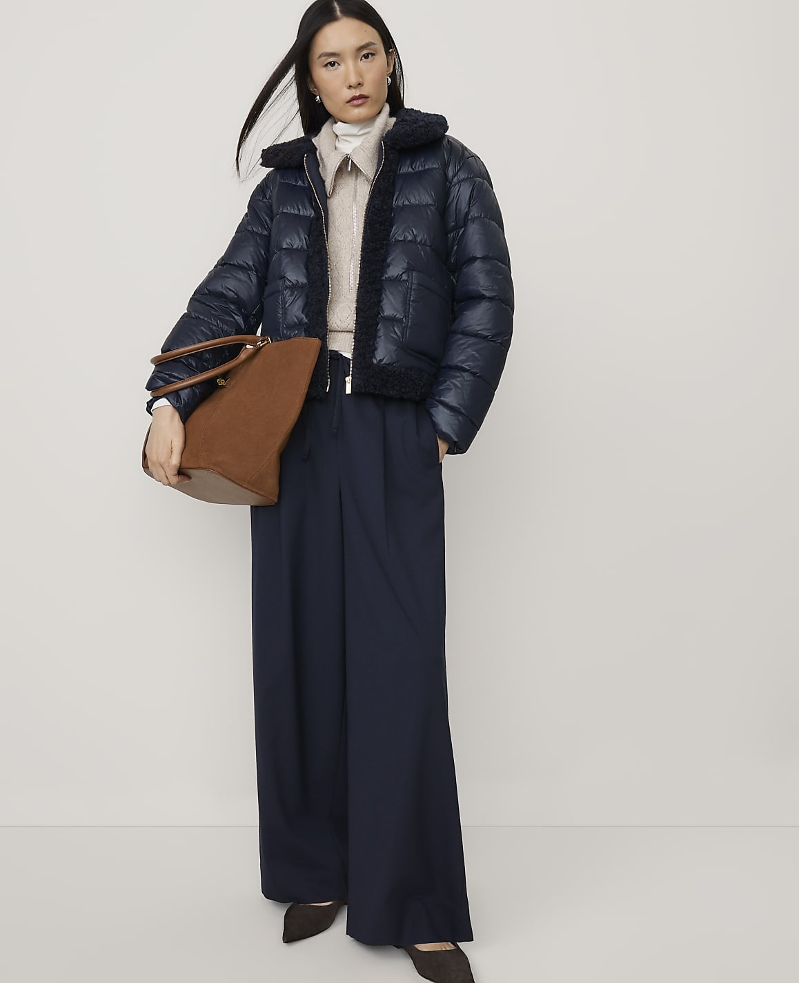 A woman wearing a navy puffer jacket and wide-leg trousers holds a brown handbag against a plain white background.