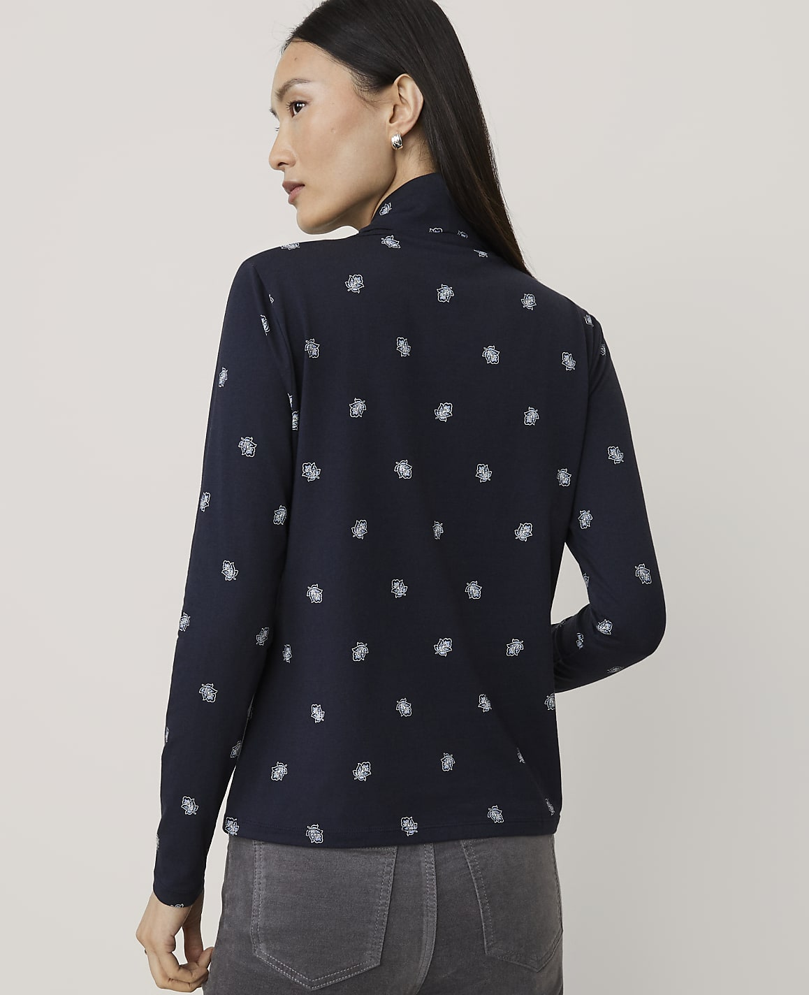 A woman wearing a navy blue patterned long-sleeve shirt and grey pants stands against a plain light grey background.