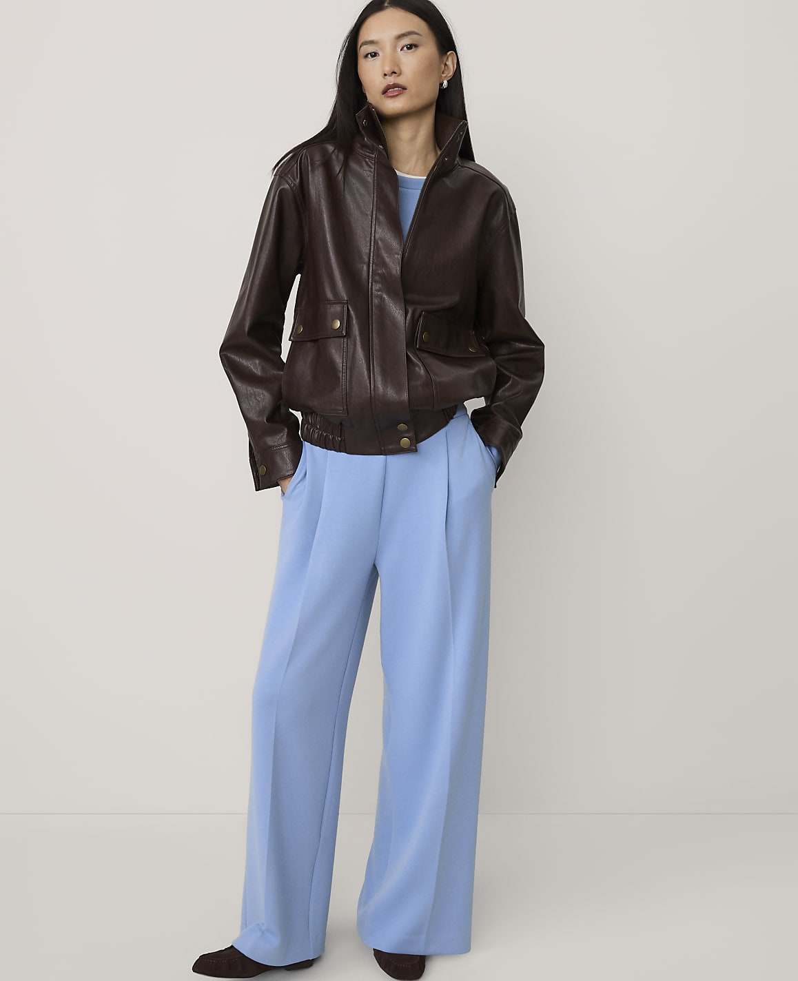 A woman wearing a brown leather jacket and wide-leg light blue trousers stands against a plain, neutral-colored background.