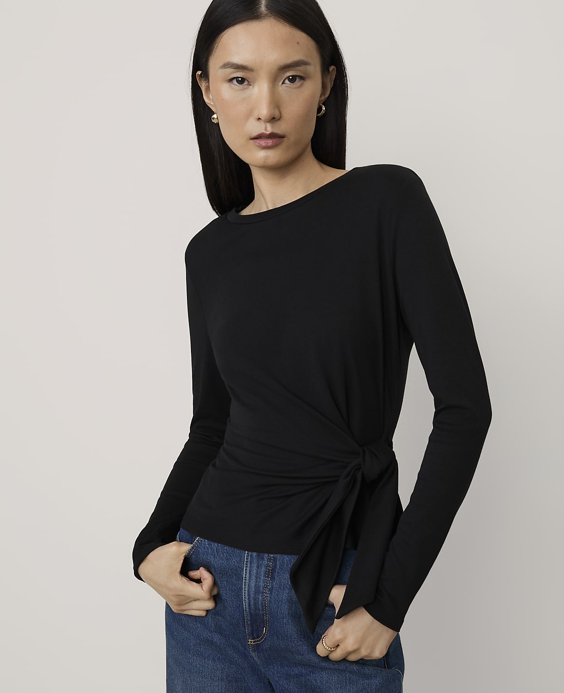 A woman wearing a black long-sleeved wrap top and blue jeans stands against a plain, light-colored background.