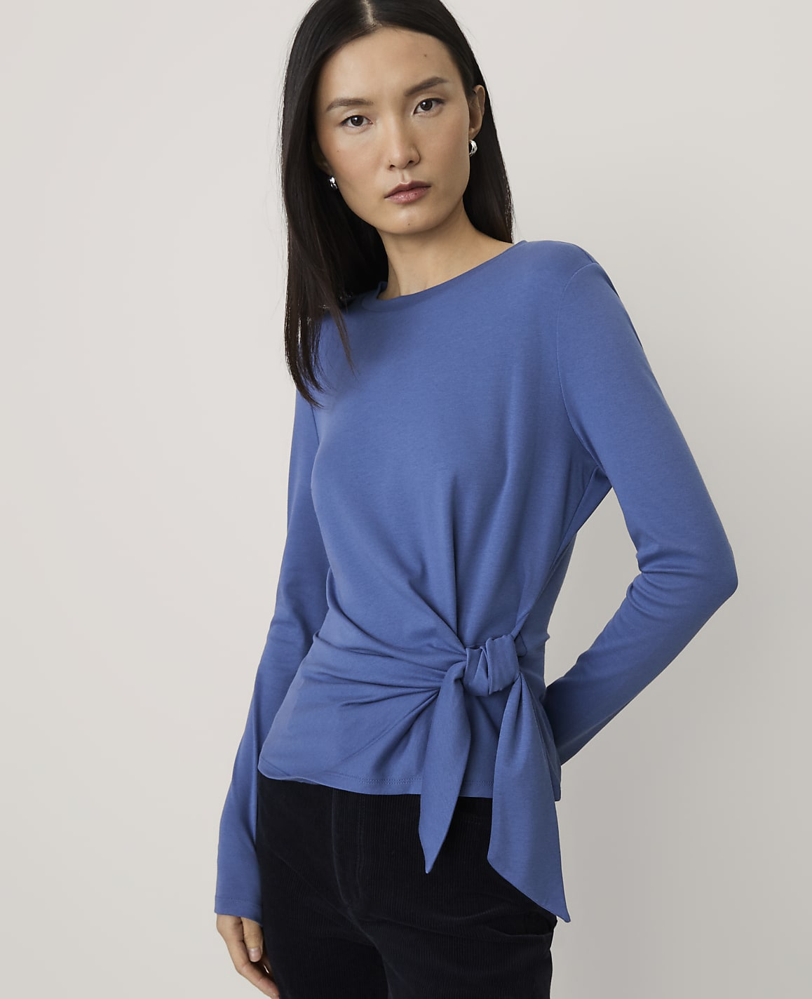 A woman wearing a blue long-sleeved wrap top and black pants stands against a plain, light-colored background.