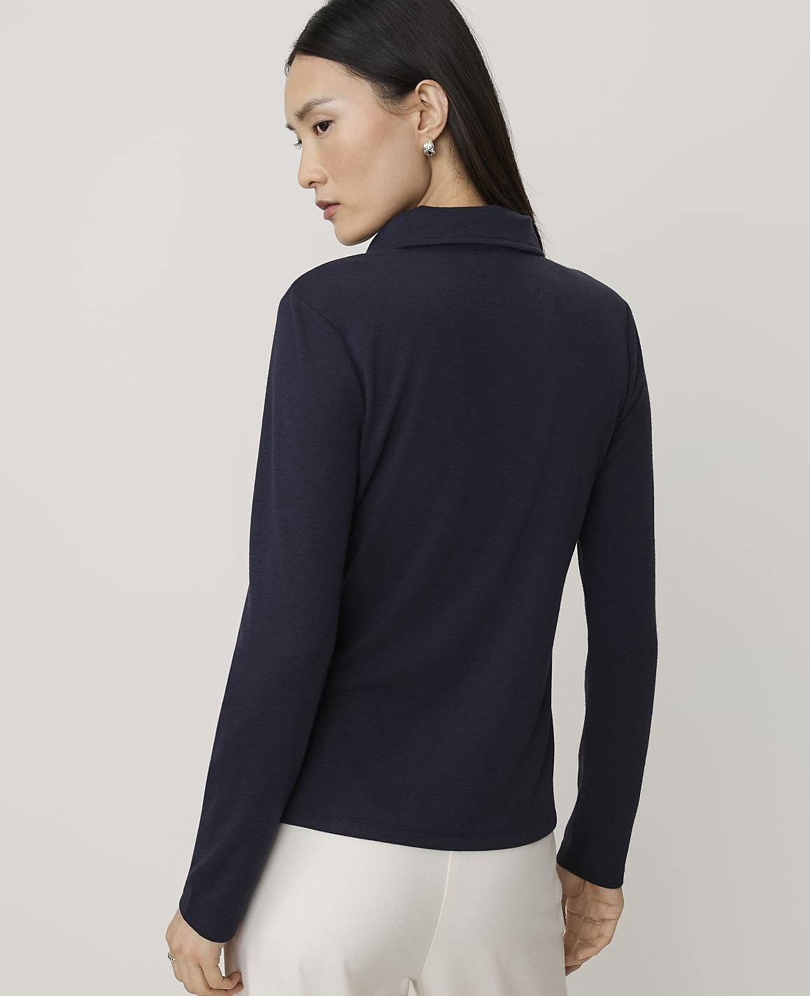A woman wearing a navy blue long-sleeved top and white trousers stands against a plain, light grey background.