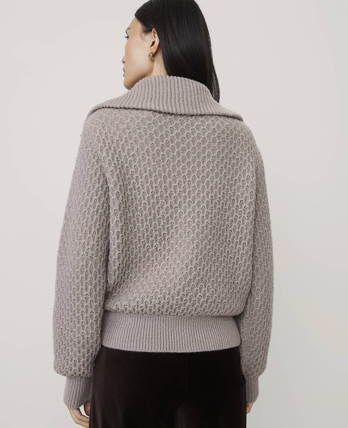 A woman wearing a textured taupe sweater and dark brown trousers stands against a plain light gray background.