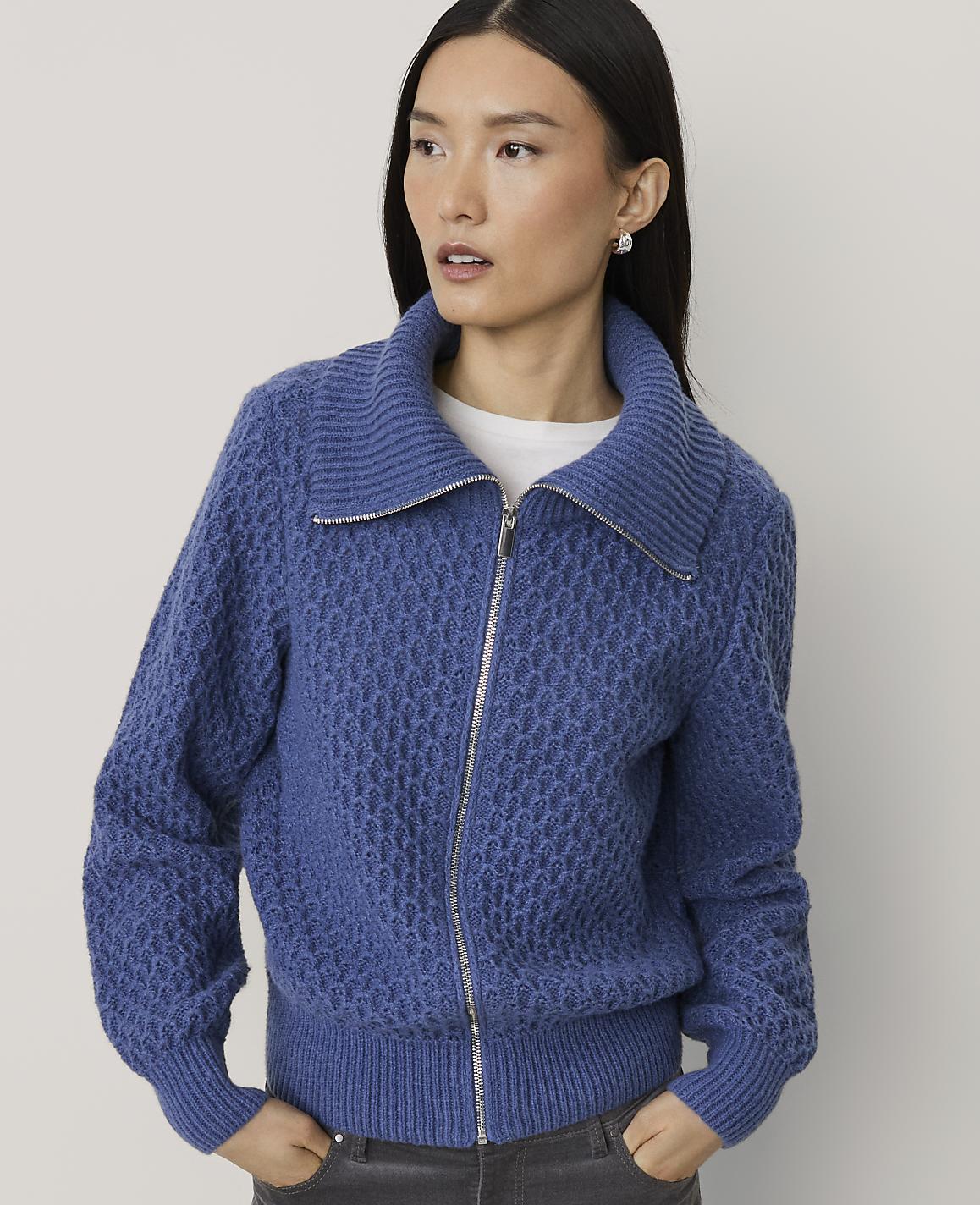 A woman wearing a blue textured zip-up jacket stands against a plain, light-colored background.