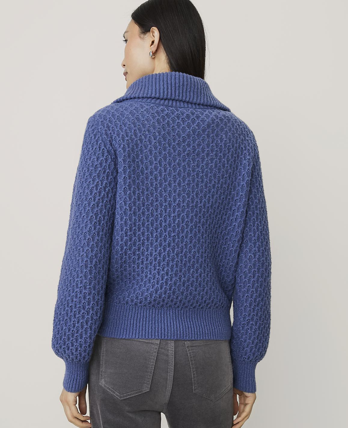 A woman wearing a blue textured knit sweater and grey pants stands against a plain light-colored background.