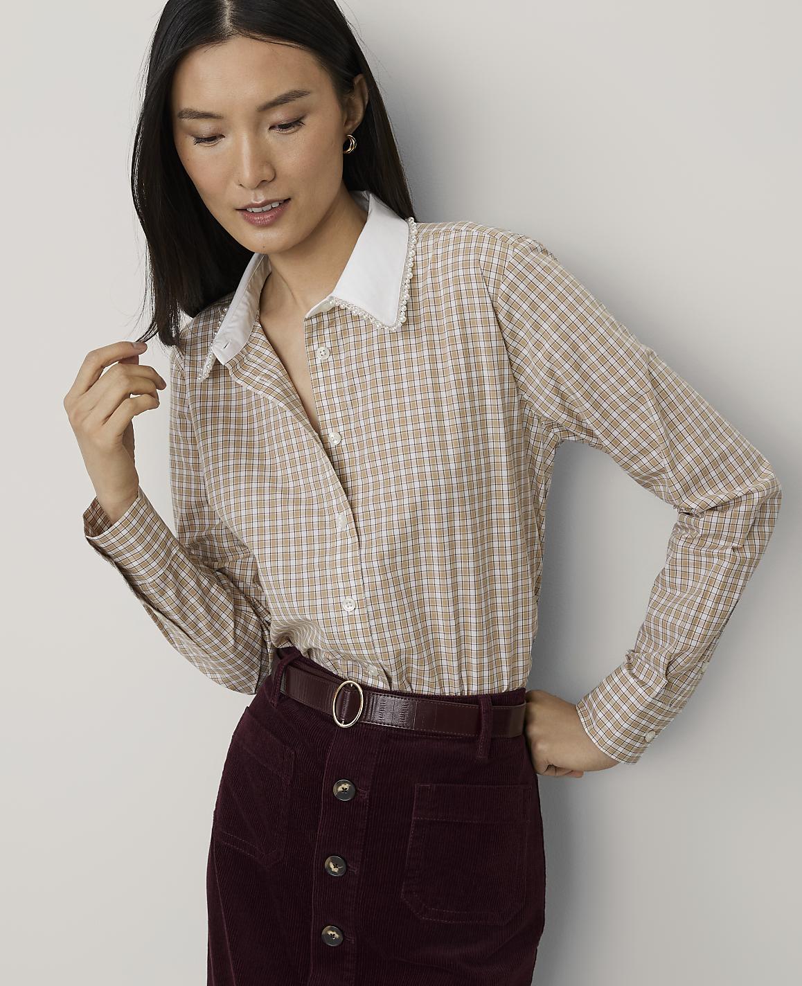 A woman wearing a tan checkered shirt and burgundy corduroy skirt stands against a plain light gray background.
