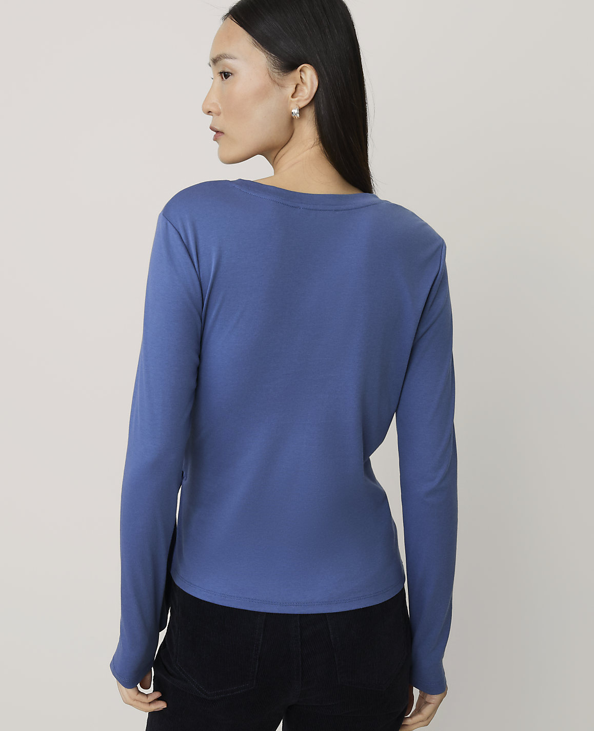 A woman wearing a blue long-sleeved shirt and black pants stands against a plain, light-colored background.