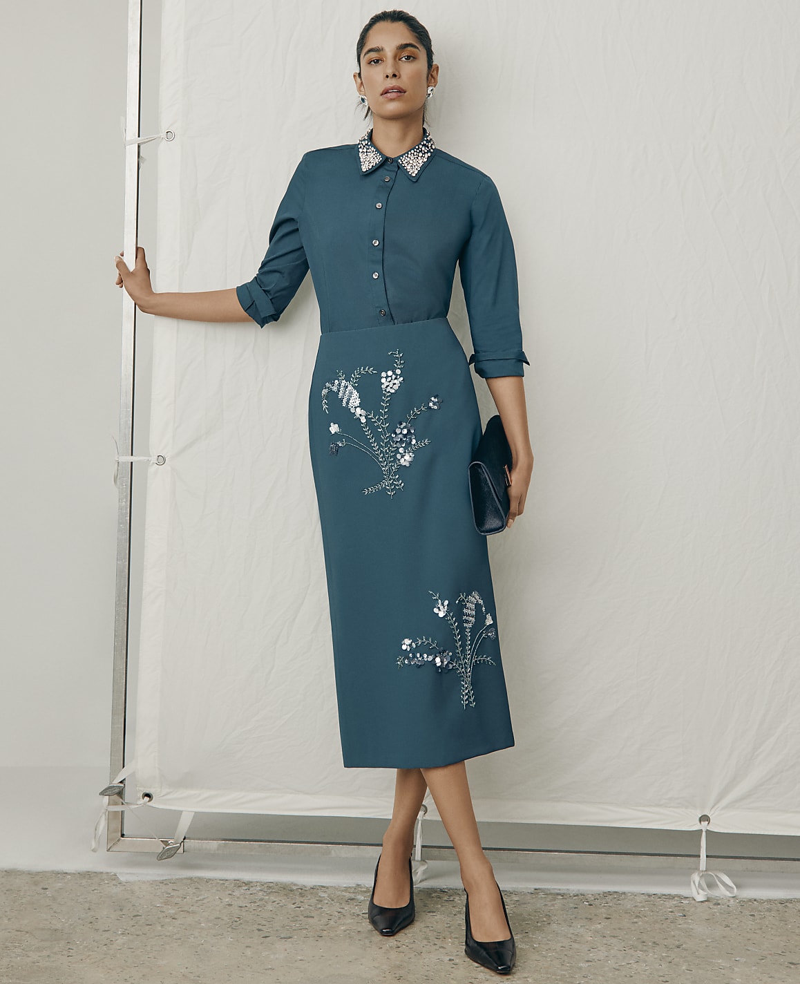 A woman wearing a teal button-down dress with floral sequins and black heels stands against a plain white fabric backdrop.