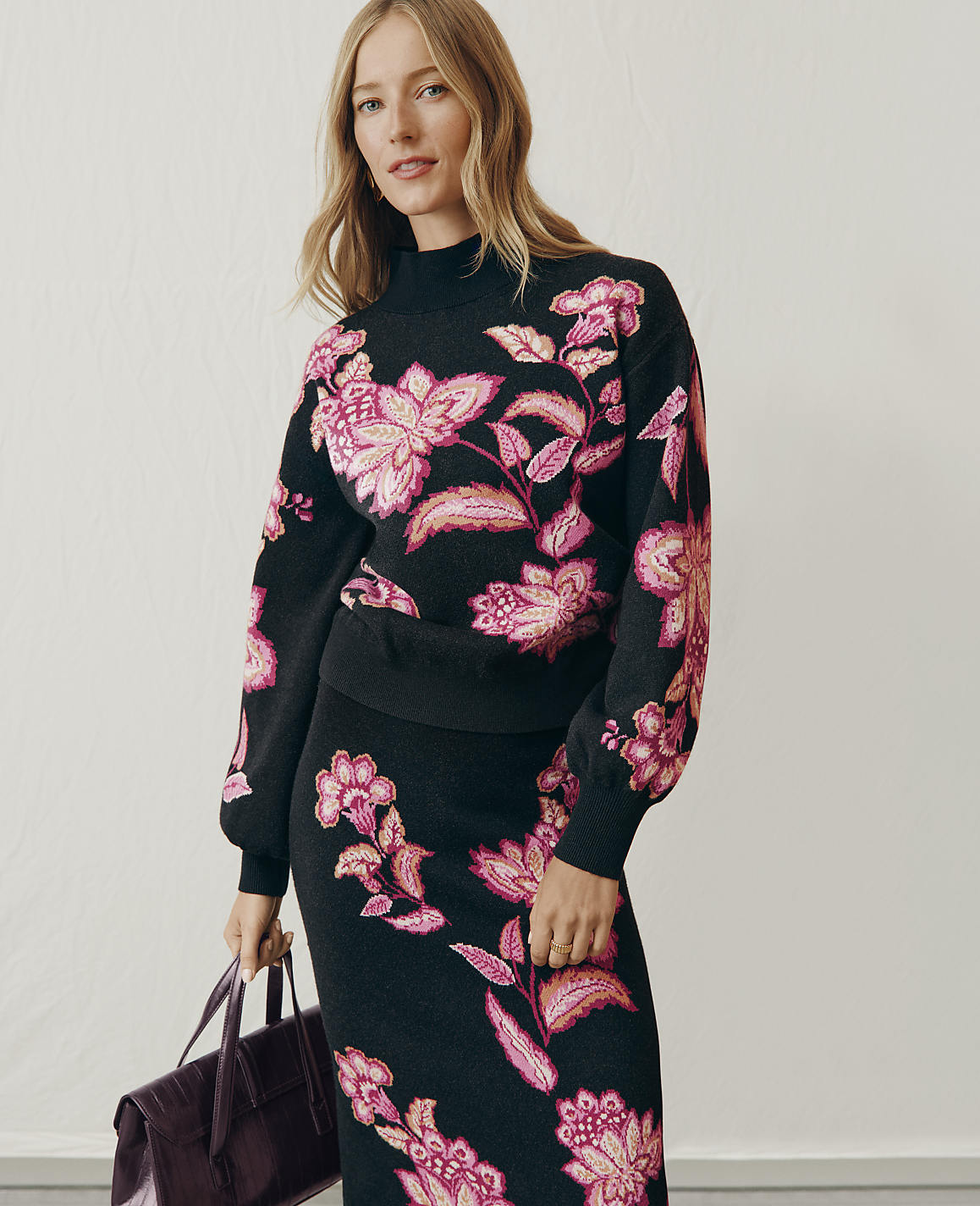 A woman wearing a black floral print knit set and holding a dark purple handbag stands against a plain white wall.