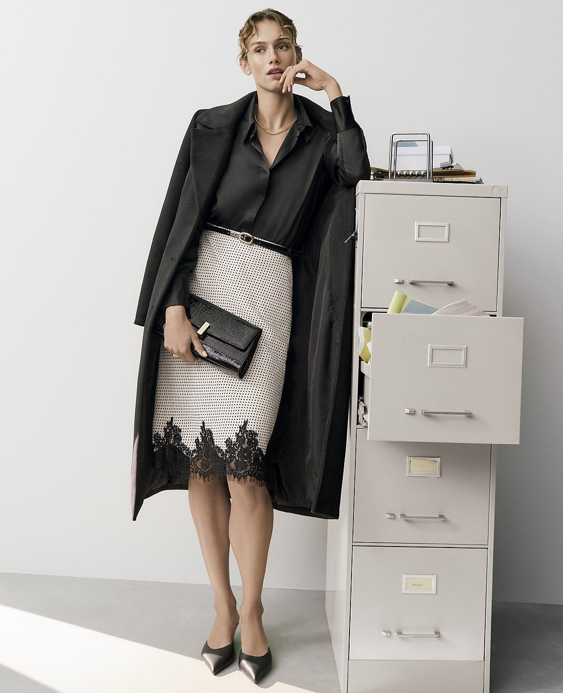 A woman in a black blouse, lace-trimmed skirt, and long coat stands next to a grey filing cabinet against a plain white background.
