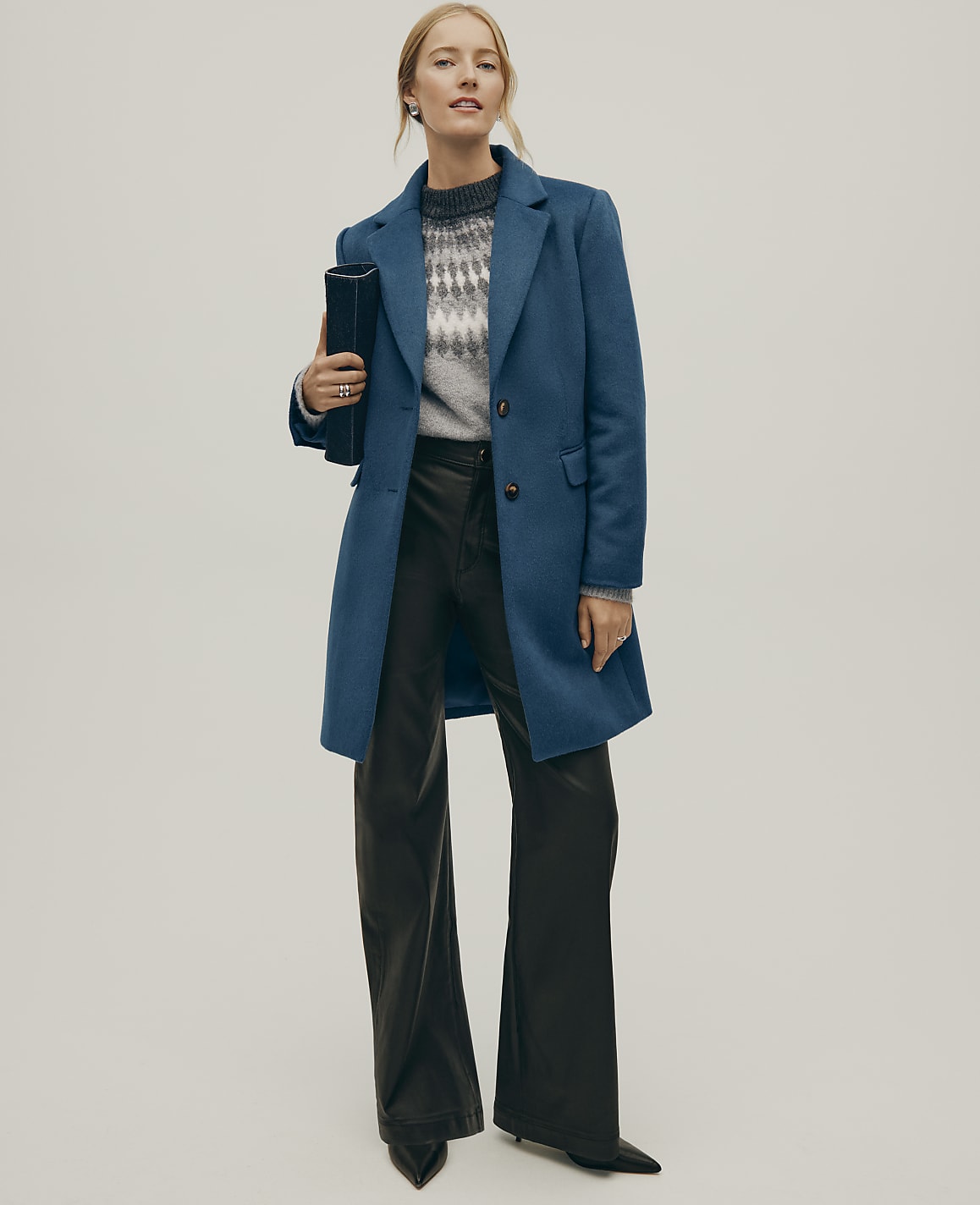 A woman wearing a blue coat, a grey patterned sweater, and black leather trousers stands against a plain, light-colored background.