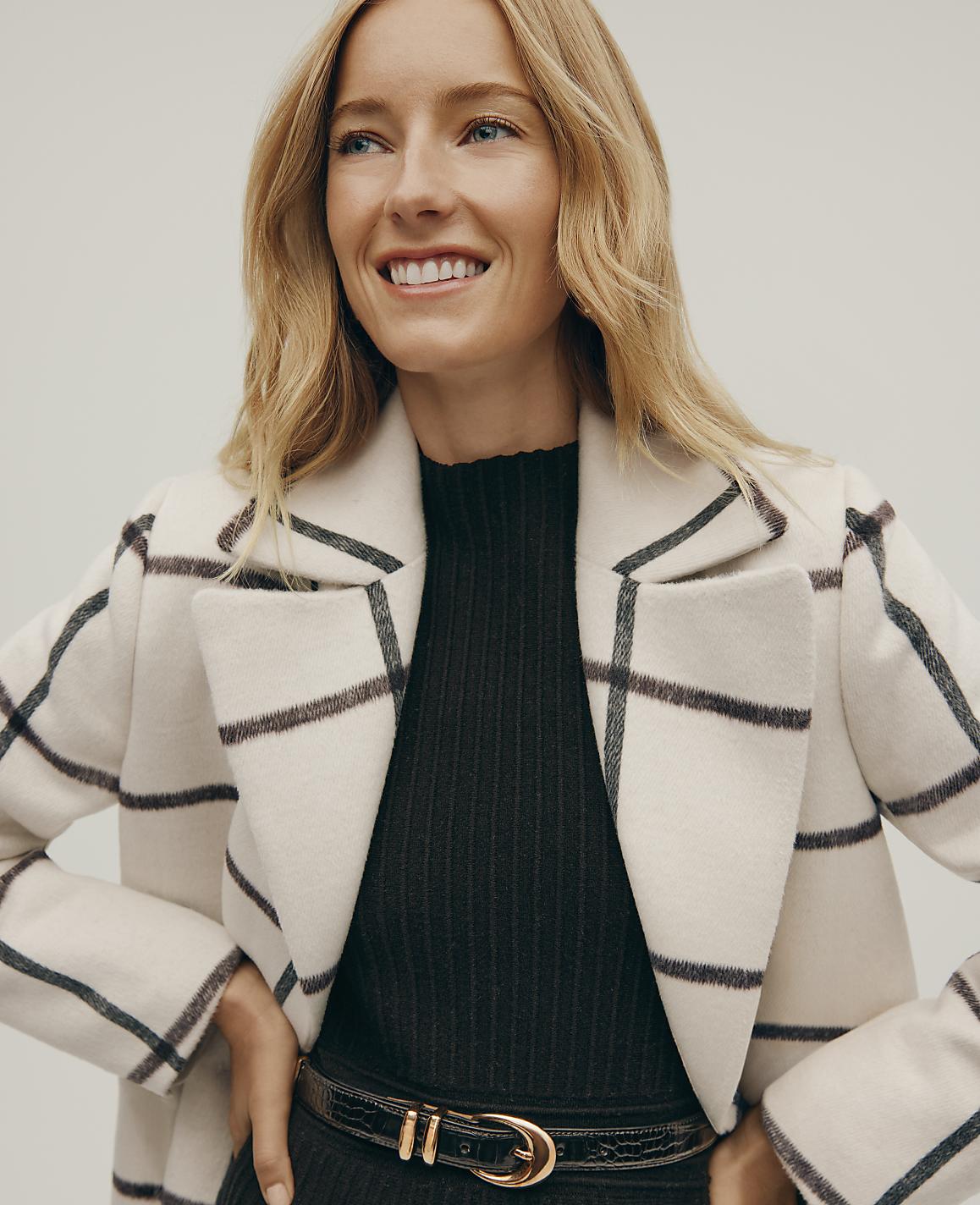A smiling blonde woman wears a white grid-patterned coat over a black ribbed top and belt against a plain light gray background.